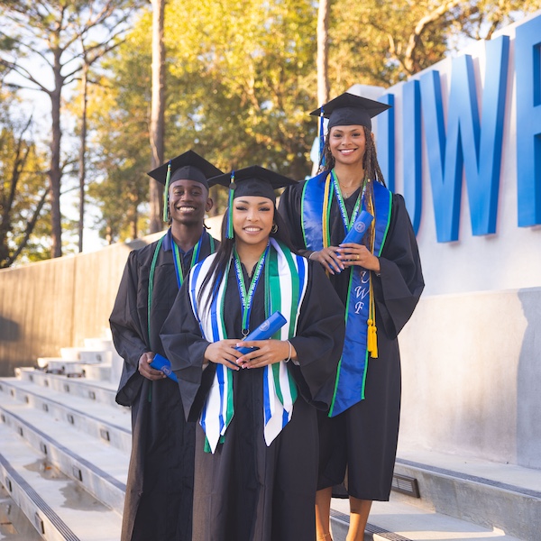 Three University of West Florida students wear black caps and gowns in front of a blue UWF sign.