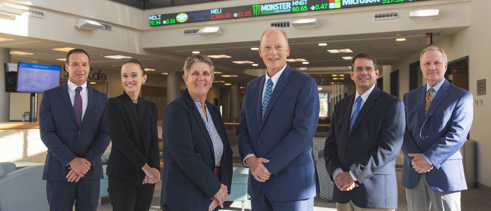 Six faculty members in suits posed in a building with a stock market ticker