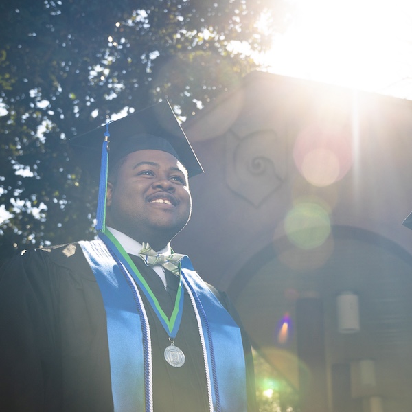 A man wears a black cap and gown and stands in front of a brick archway.