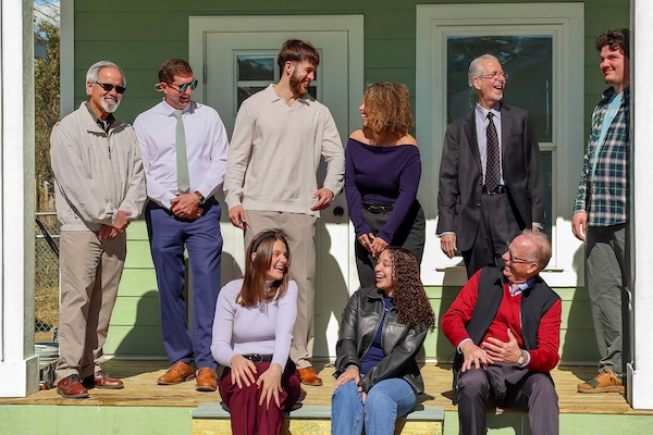 Three people sitting on steps and six people standing on porch of tiny home