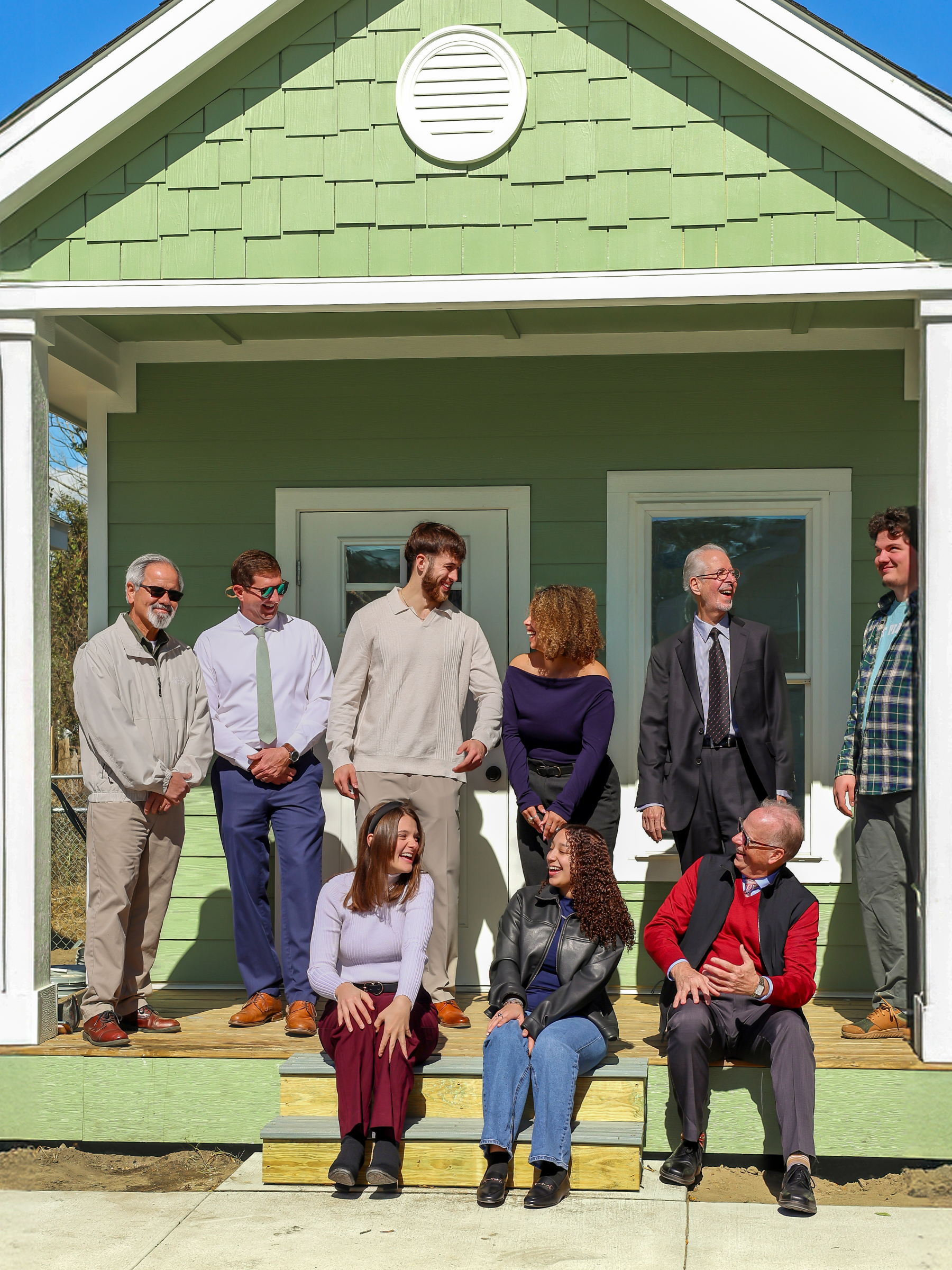 Three people sitting on steps and six people standing on porch of tiny home