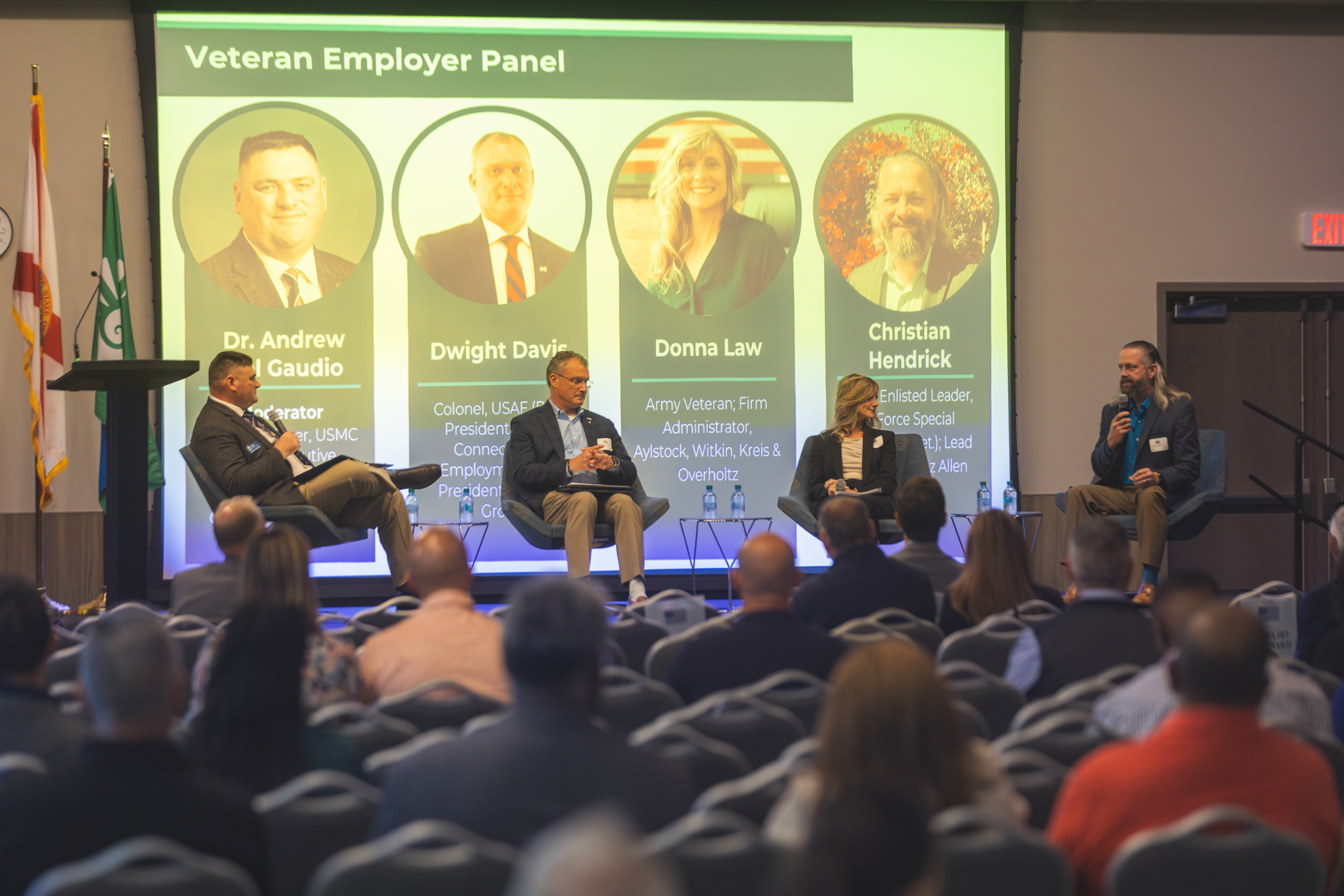 Four speakers sitting in chairs on a stage during a conference in front of a large screen displaying Veteran Employer Panel showcasing the speakers.