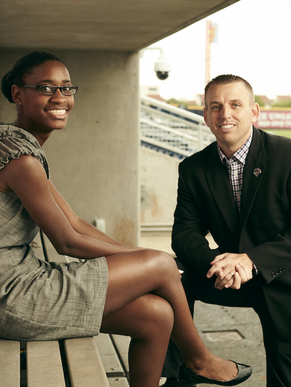 A business mentor and student both in professional attire sitting in a dugout at a baseball stadium with the baseball field visible in the background.