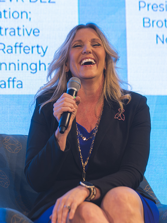 Conference speaker speaking into a microphone while sitting on a stage chair in front of a large display screen.