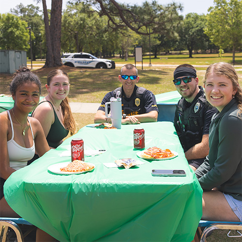 Students and Police officers eating pizza at a table, smiling at the camera