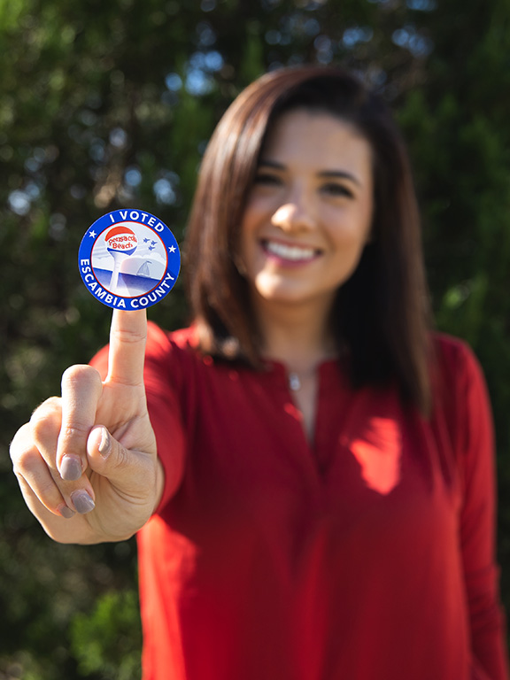 Escambia County voter smiling at camera with a sticker on their index finger that has a graphic of the Pensacola Beach water tower, sailboat and three jets encircled by 