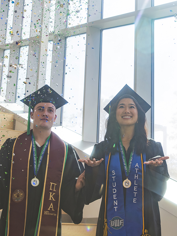 Two UWF graduates in cap and gown attire throwing confetti