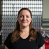 Jennifer Eaton in a black shirt poses for an indoor upper body portrait in front of a large stack of silver, unlabeled drinking cans.