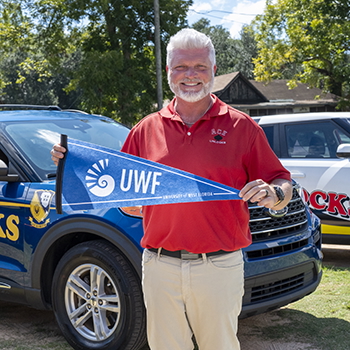 Christopher Roney outdoors holding a University of West Florida pennant while standing in front of two SUVs.