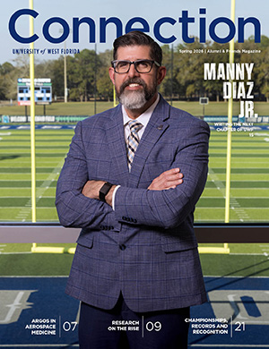Cover of the Spring 2026 Connection magazine featuring UWF President Manny Diaz posing with his arms crossed standing indoors in front of a window with the UWF football field in the background.