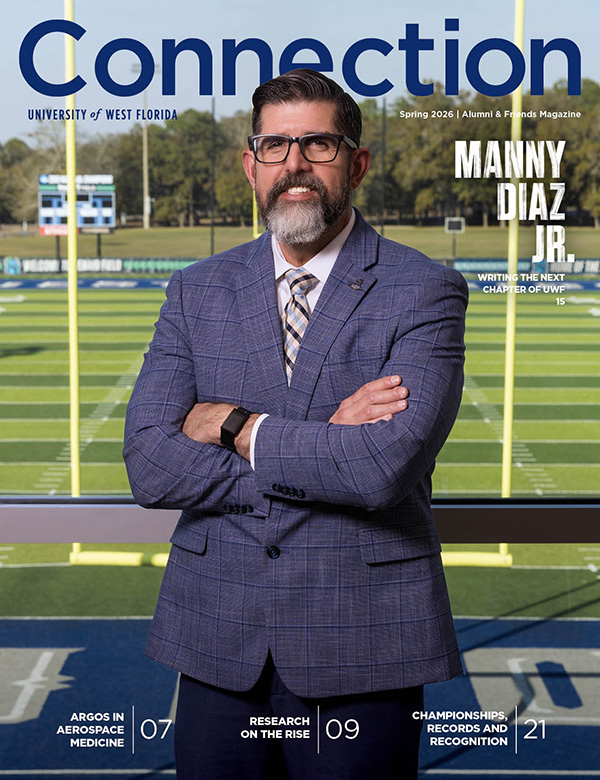 Cover of the Spring 2026 Connection magazine featuring UWF President Manny Diaz posing with his arms crossed standing indoors in front of a window with the UWF football field in the background.