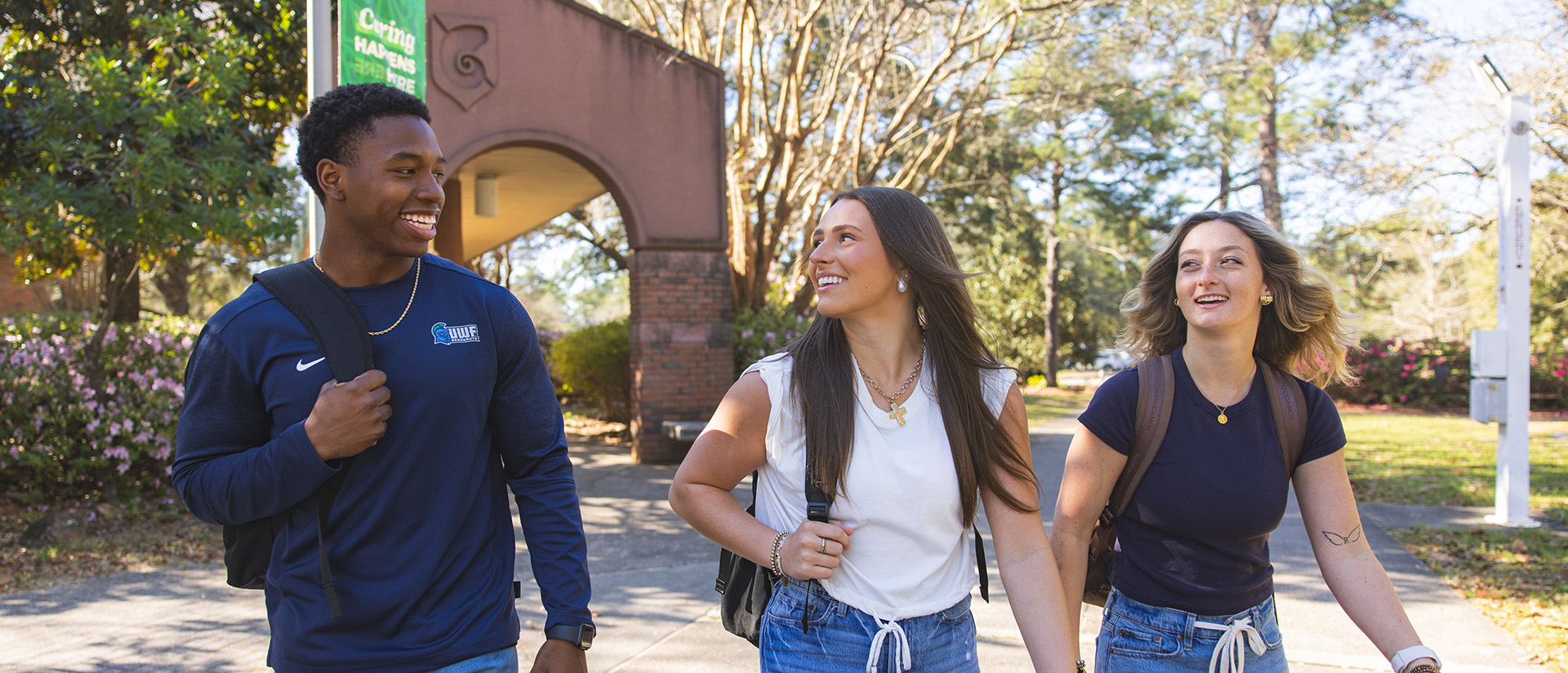 Three students walking on a sidewalk outdoors in a horizontal row with a brick archway and trees in the background.