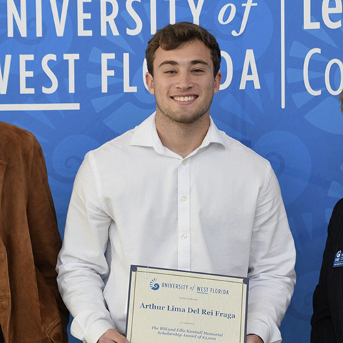 Arthur Lima d'El-Rei Fraga holding an award certificate with his name while standing in front of a University of West Florida step-and-repeat backdrop.