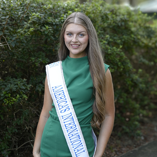 Bailey Scheel standing outdoors in front of a bush wearing a green outfit with a sash that states America's International.