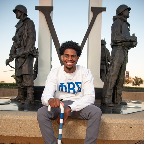 Chris Meriweather wearing a long sleeve shirt with Greek letters while holding a blue and white striped cane and sitting in front of a monument with a large V and three statues of soldiers.