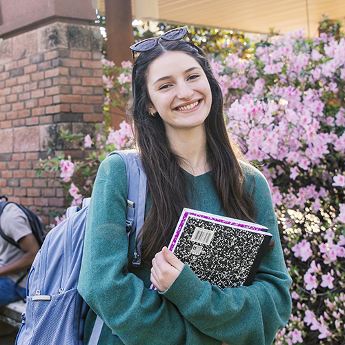 Elisabetta Savini standing outdoors smiling at the camera wearing a backpack and holding two notebooks against her chest while standing in front of a pink flowering bush.