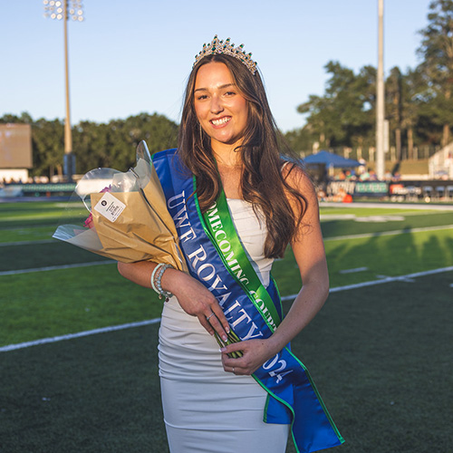 Emma Morgan outdoors on a football field in a homecoming dress with a crown on her head and two homecoming sashes wrapped around her while she is holding a bouquet of flowers.