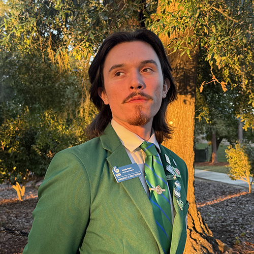 Jacob Hyer outdoors standing in front of trees looking off into the distance while wearing a green suit and tie with a UWF name badge and 8 lapel pins.