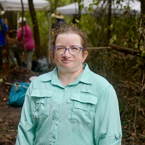 Jennifer Melcher smiles in a forested setting wearing a light blue long-sleeve shirt with activity of an archeological dig in the background.