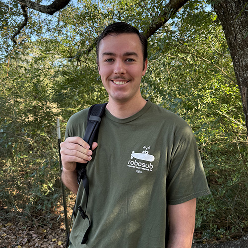 Jonathan Lavoie smiling at the camera while wearing a backpack and standing outdoors in front of a tree and bushes.