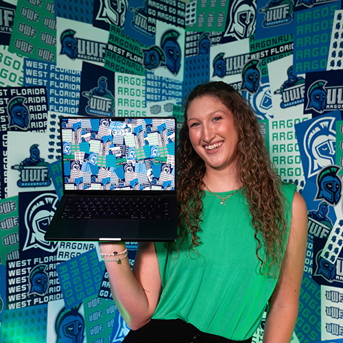 Jordan Taylor smiling at the camera while holding a laptop with the screen facing that camera displaying a UWF pattern theme background which is also displayed on the wall behind her.