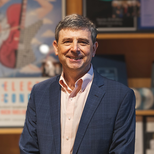 Joseph Spaniola in professional attire standing indoors in front of a wall displaying various musical awards and art pieces.