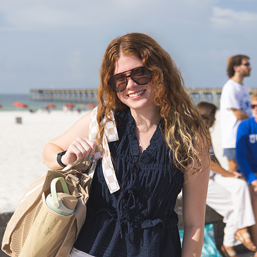 Kyleigh Rawlins smiling at the camera while standing in front of the sand at the beach with the water and a fishing pier in the background.