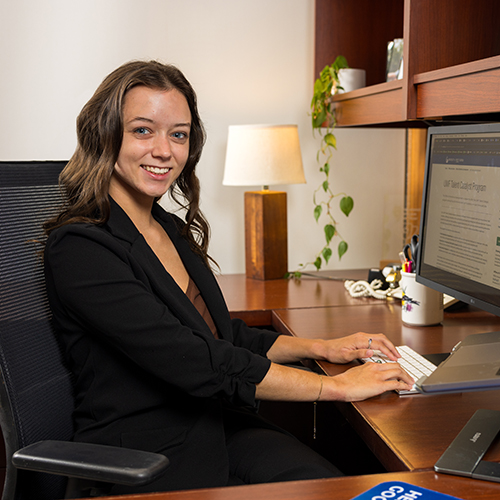 Savannah Guilford smiling at the camera while sitting in an office chair and typing on a keyboard at a large desk in an office space.