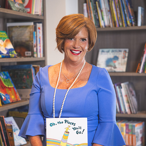 Susan James smiling at the camera while standing in a library while holding Oh, the Places You'll Go! by Dr. Seuss.