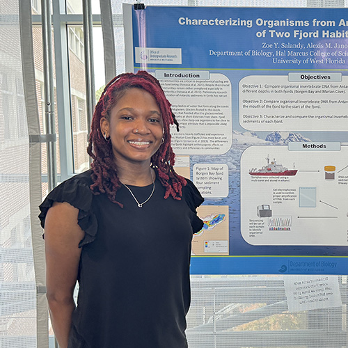 Zoe Salandy smiling at the camera while standing in front of a science project display board titled Characterizing Organisms from Antarctic Sediments of Two Fjord Habitats.