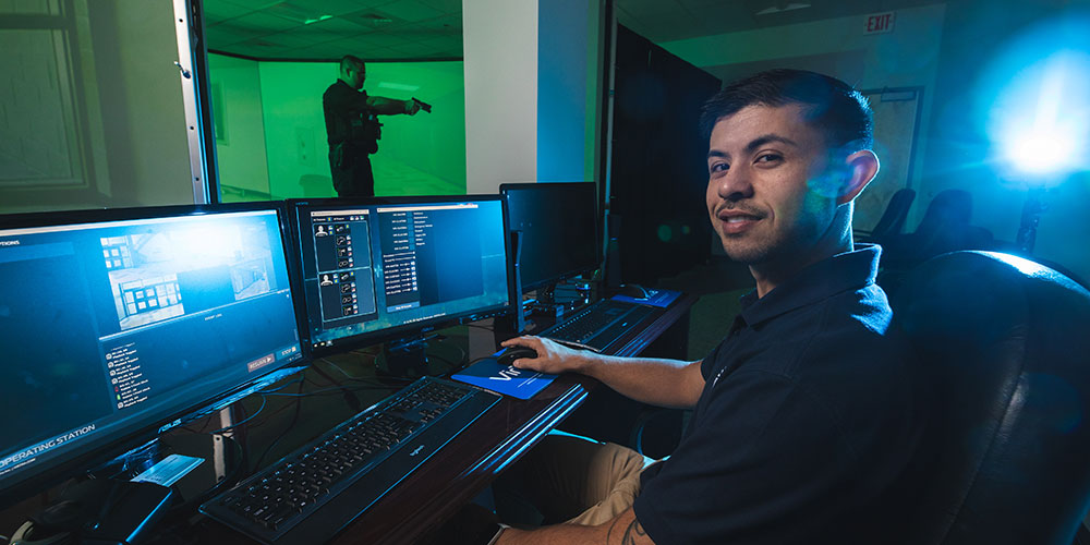 Four students sitting in a row at a table in a classroom in the foreground with four other students sitting at tables in the background.