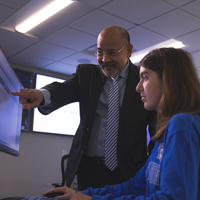 A cybersecurity faculty pointing at a computer monitor that a student is sitting in front of in a classroom.