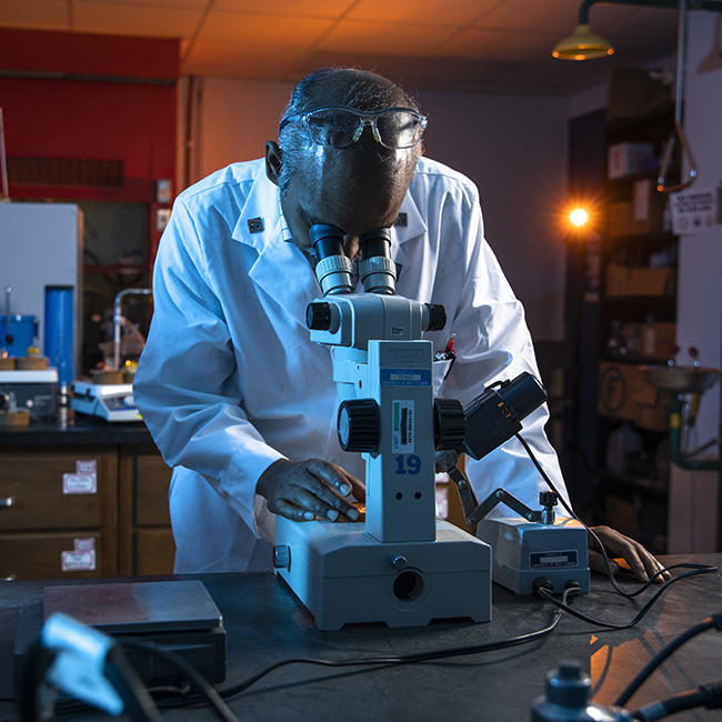 A chemistry professor wearing lab attire while looking through microscope equipment in a lab classroom.