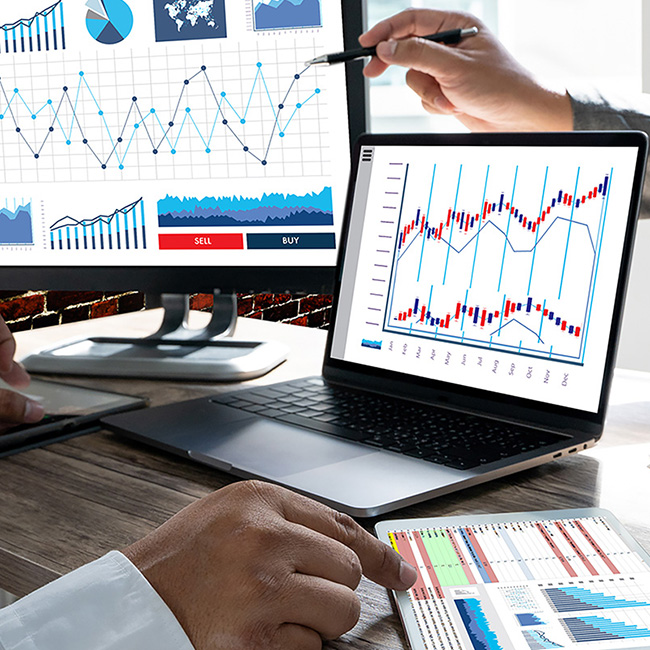 Hands of two people at a desk using a laptop, monitor, and printout displaying various data analysis charts and figures.