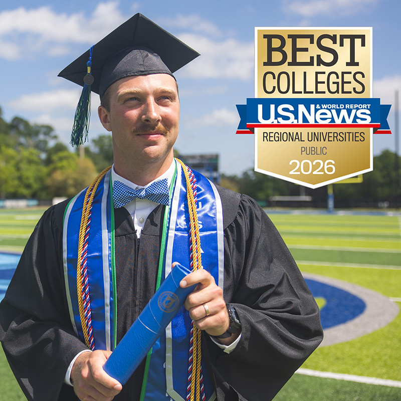 A student in commencement cap and gown attire holding a diploma tube standing on a football field with a U.S. News and World Report Best Colleges Regional Universities Public 2026 graphic at the top right of the image.