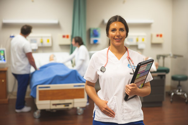 Nursing student stands in front of other nursing students who interact with a simulated patient.
