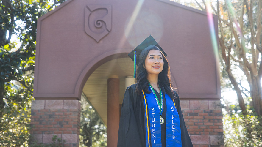 UWF graduate in cap and gown attire in front of the Cantor al Sol archway on the Pensacola campus.