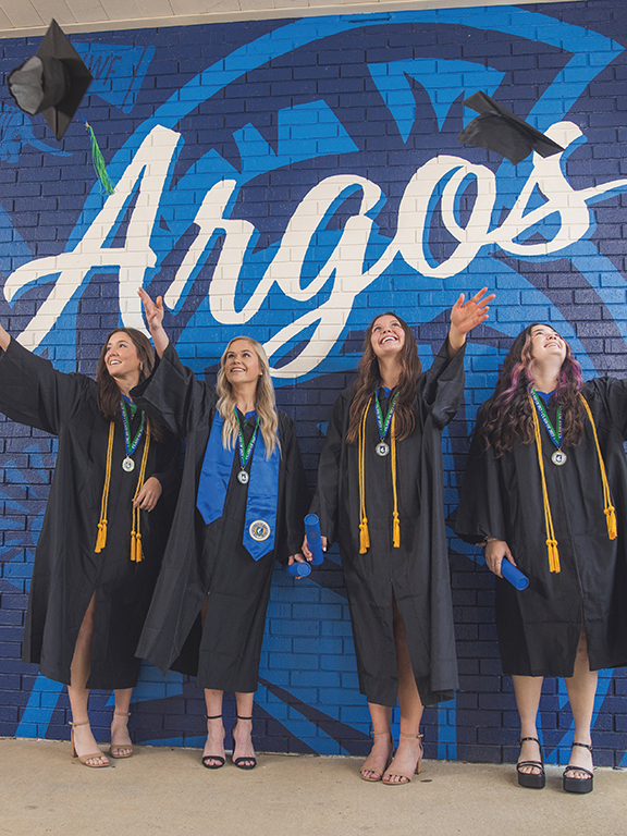 Four UWF graduates in cap and gown attire in front of the Go Argos painted mural on the UWF Pensacola campus.