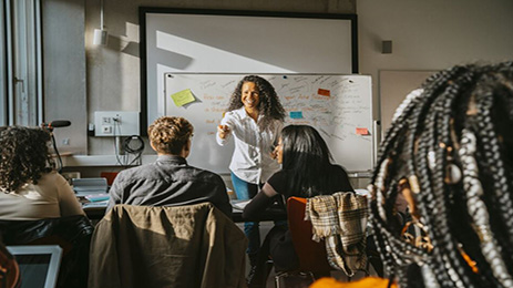 A professor in a classroom setting gestures enthusiastically while teaching in a classroom filled with students seated at desks. A whiteboard with written notes and sticky notes is in the background.