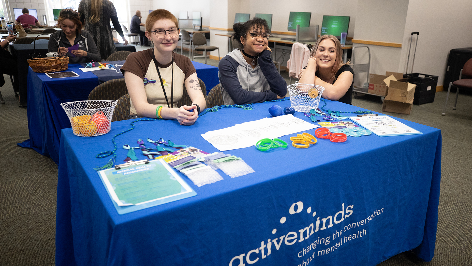 Mental health awareness booth with pamphlets, wristbands, and volunteers at a library event. sitting at an exhibit table.