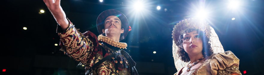 Two theatre students dressed in Shakespeare costumes look toward the camera as spotlights shine behind them.