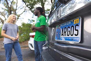 Two students conversing outdoors behind a vehicle that has an Argos license plate.