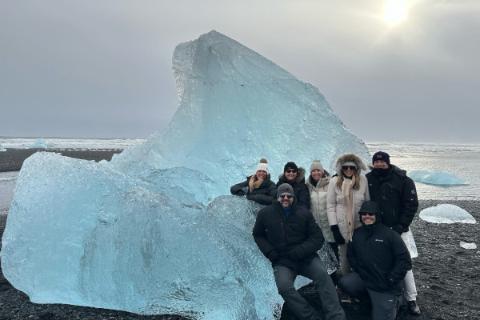 A group of travelers in heavy jackets pose next to an iceberg that is much taller than all of them.
