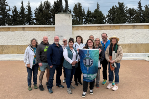 The UWF group holds a UWF flag in front of the monument to the ancient Battle of Thermopylae