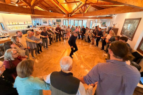 A Greek dance teacher stands in the middle of a dance circle and demonstrates a traditional Greek dance.