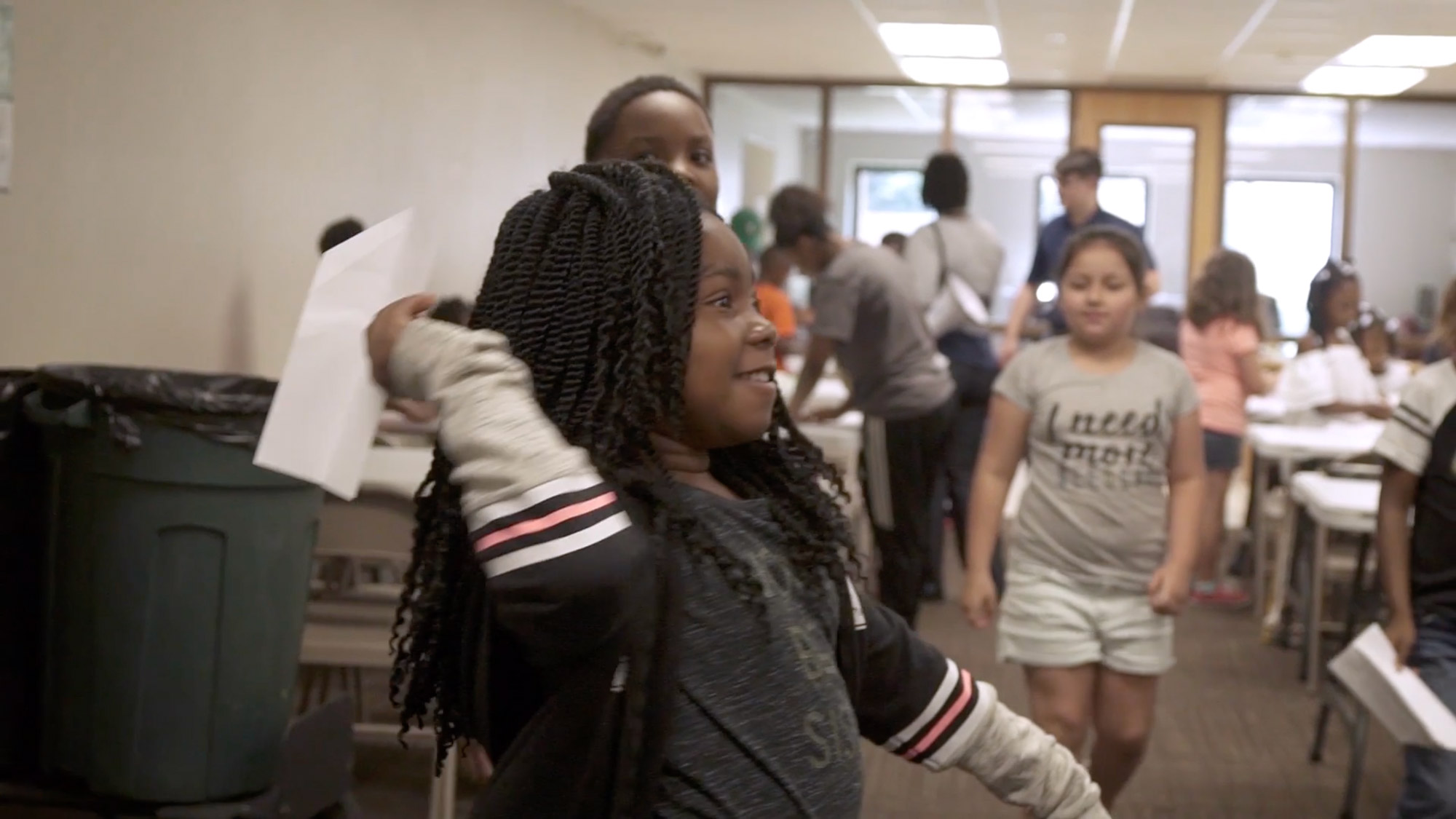 A young girl throws a paper airplane in a room full of other young people