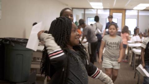 A young girl throws a paper airplane in a room full of other young people