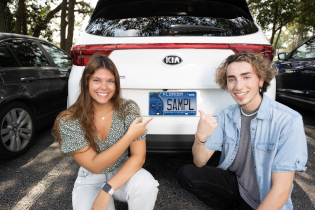 two students kneel at the trunk of a car with the Argos license plate