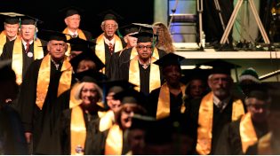 Members of the first graduating class process into the 2017 winter commencement ceremony wearing black regalia and a golden sash.