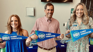 Three people are standing in front of a table holding University of West Florida Alumni Association pennants.